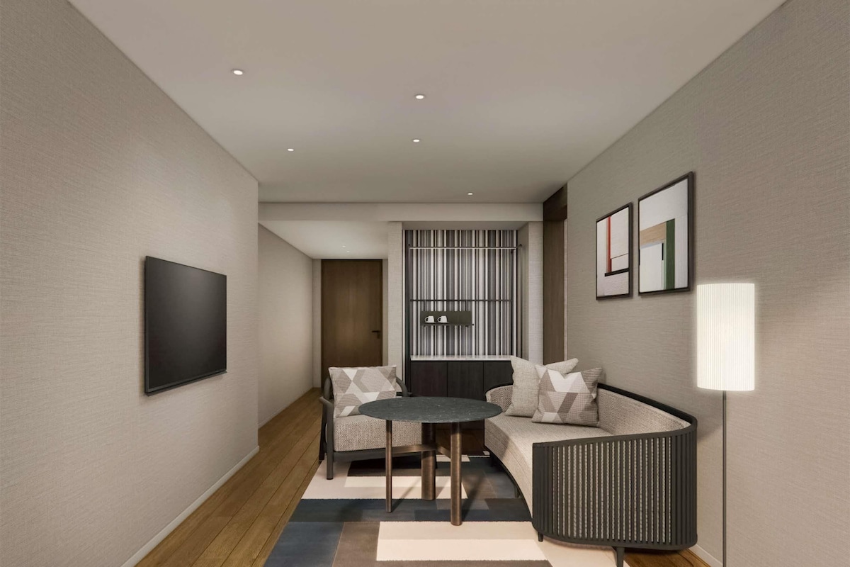A modern hotel room with a sofa, a small round table, a floor lamp, and a TV mounted on the wall.