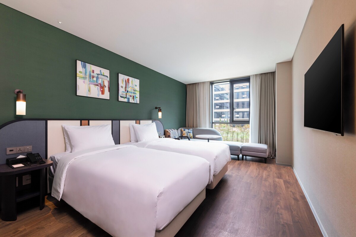Habitación de hotel con dos camas, un escritorio, una silla, un televisor y una obra de arte en la pared.