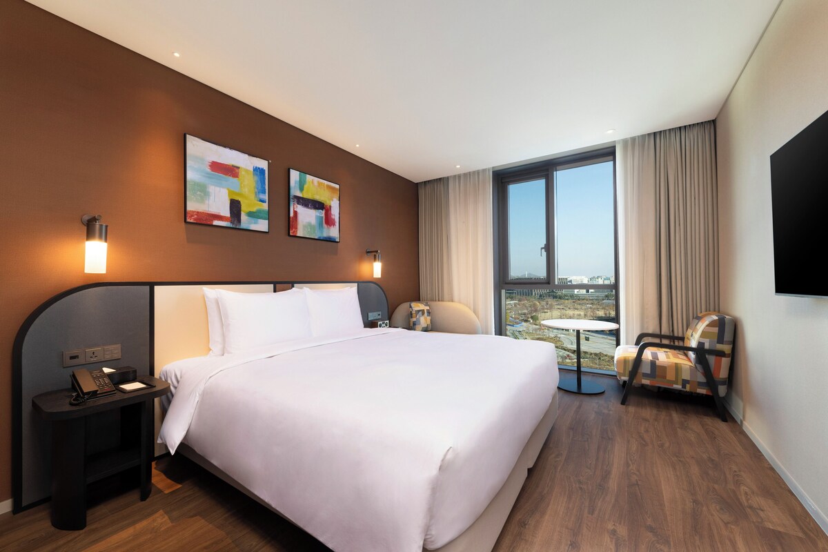 A modern hotel room with a large bed, a desk with a phone, a chair, and a view of the city through large windows.