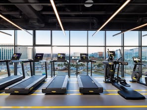 A gym with treadmills, elliptical machines, and exercise bikes, featuring large windows and a city view.