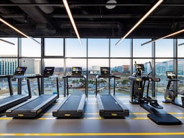A gym with treadmills, elliptical machines, and exercise bikes, featuring large windows and a city view.