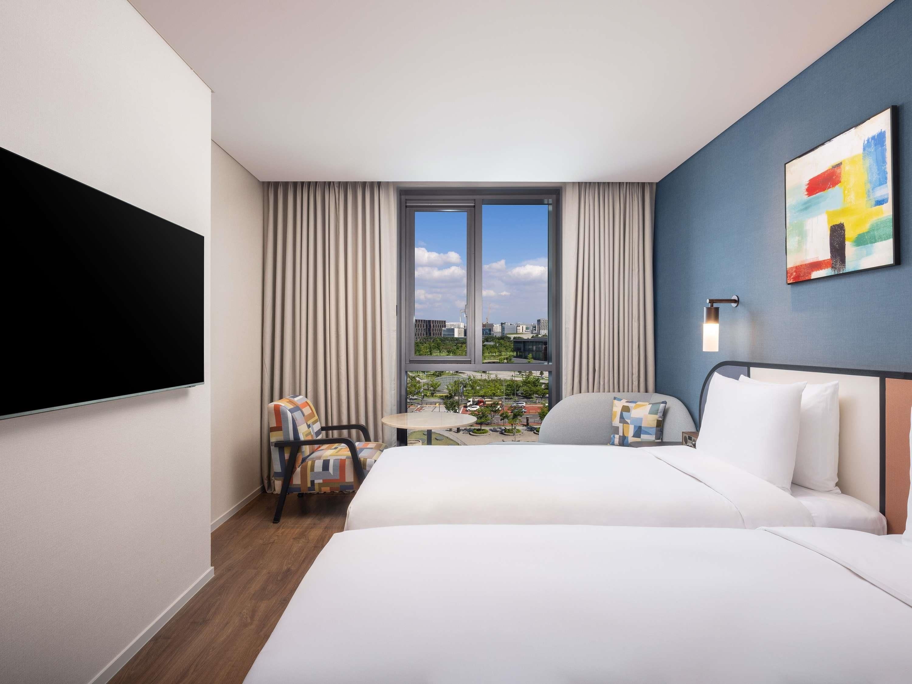 A hotel room with two beds, a TV, a small table, a chair, and a view of the cityscape.