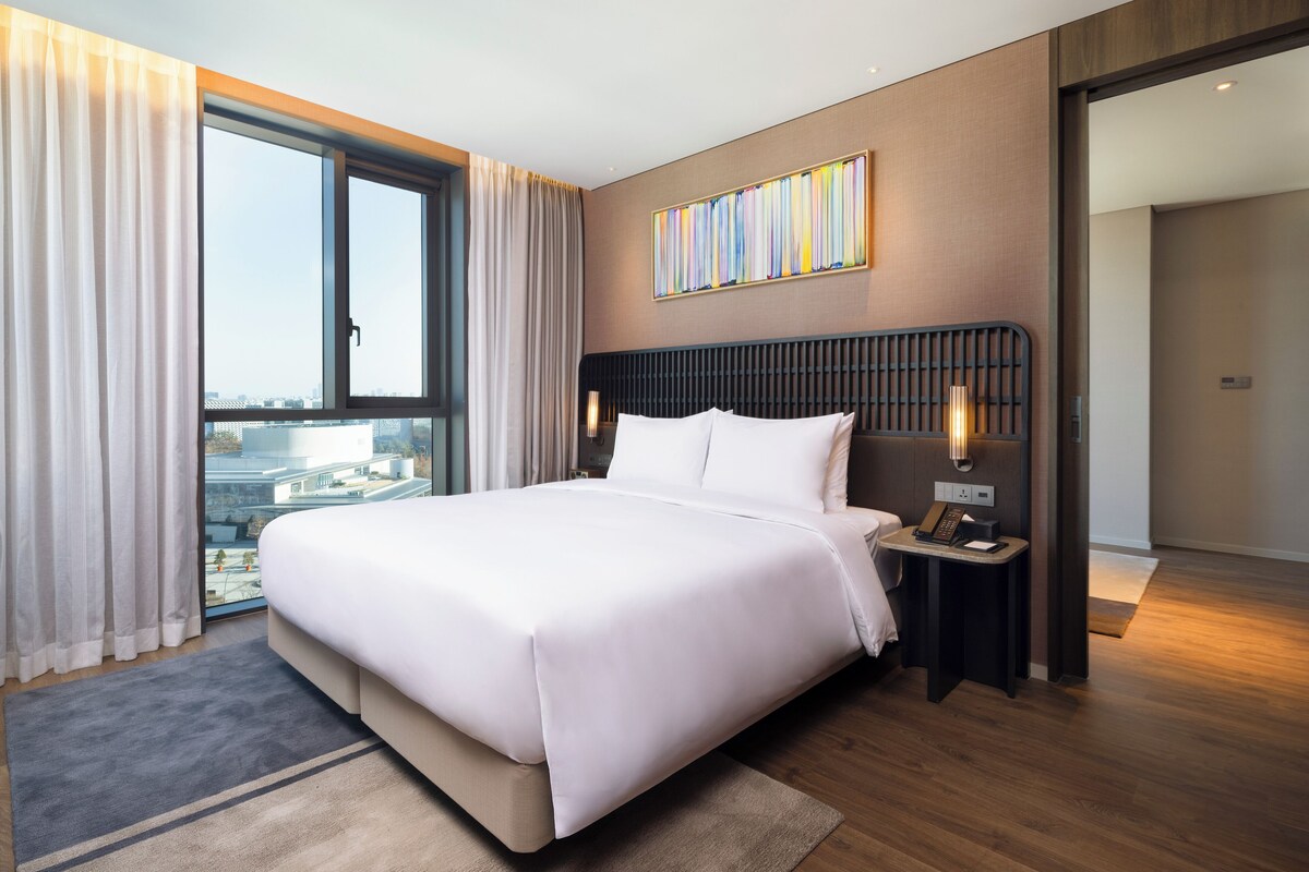 Habitación de hotel moderna con una cama grande, vistas a la ciudad, una mesita de noche con teléfono y un amplio ventanal con cortinas.