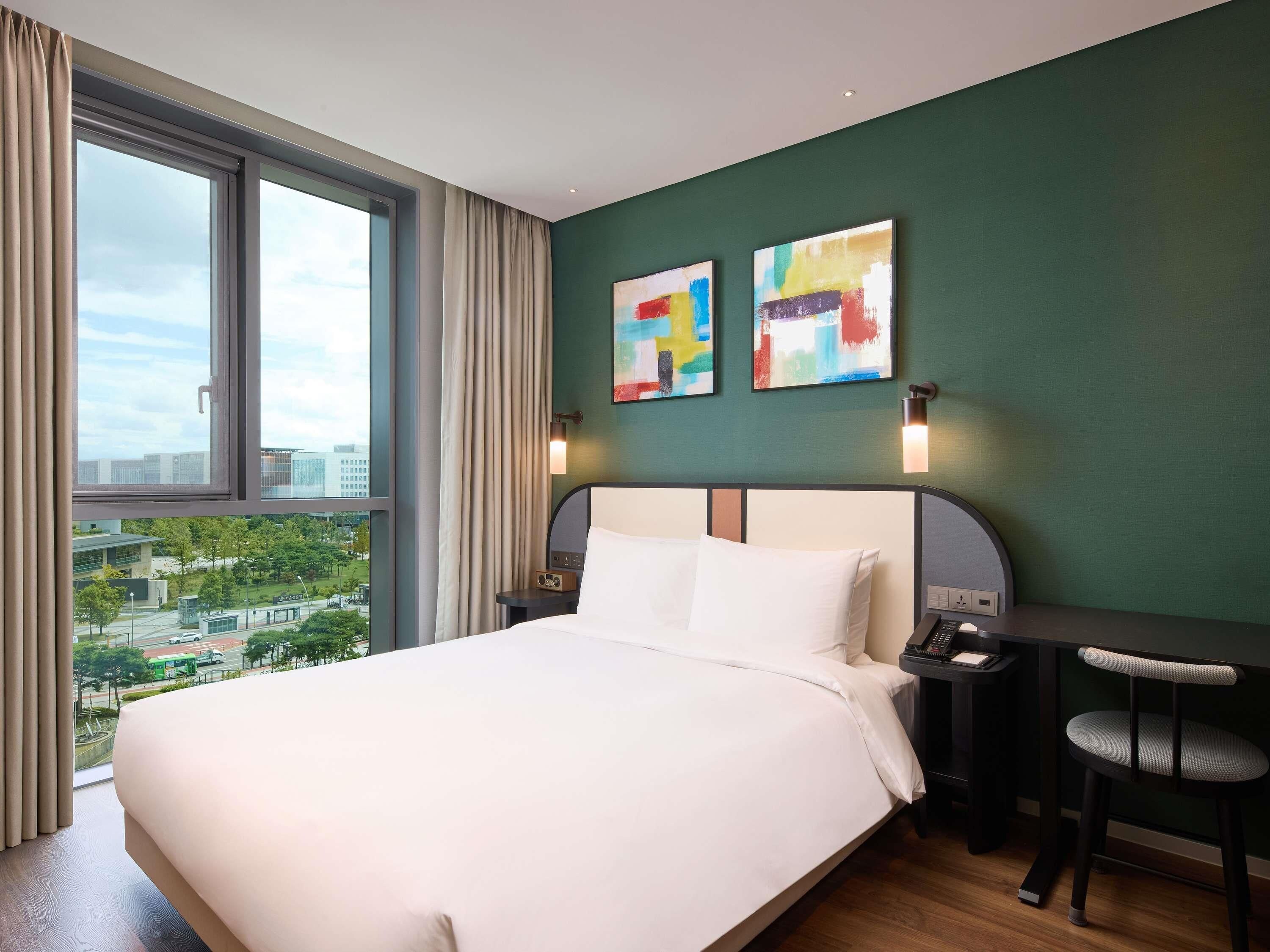 A hotel room with a large bed, a desk, two chairs, a view of the city, and abstract art on the wall.