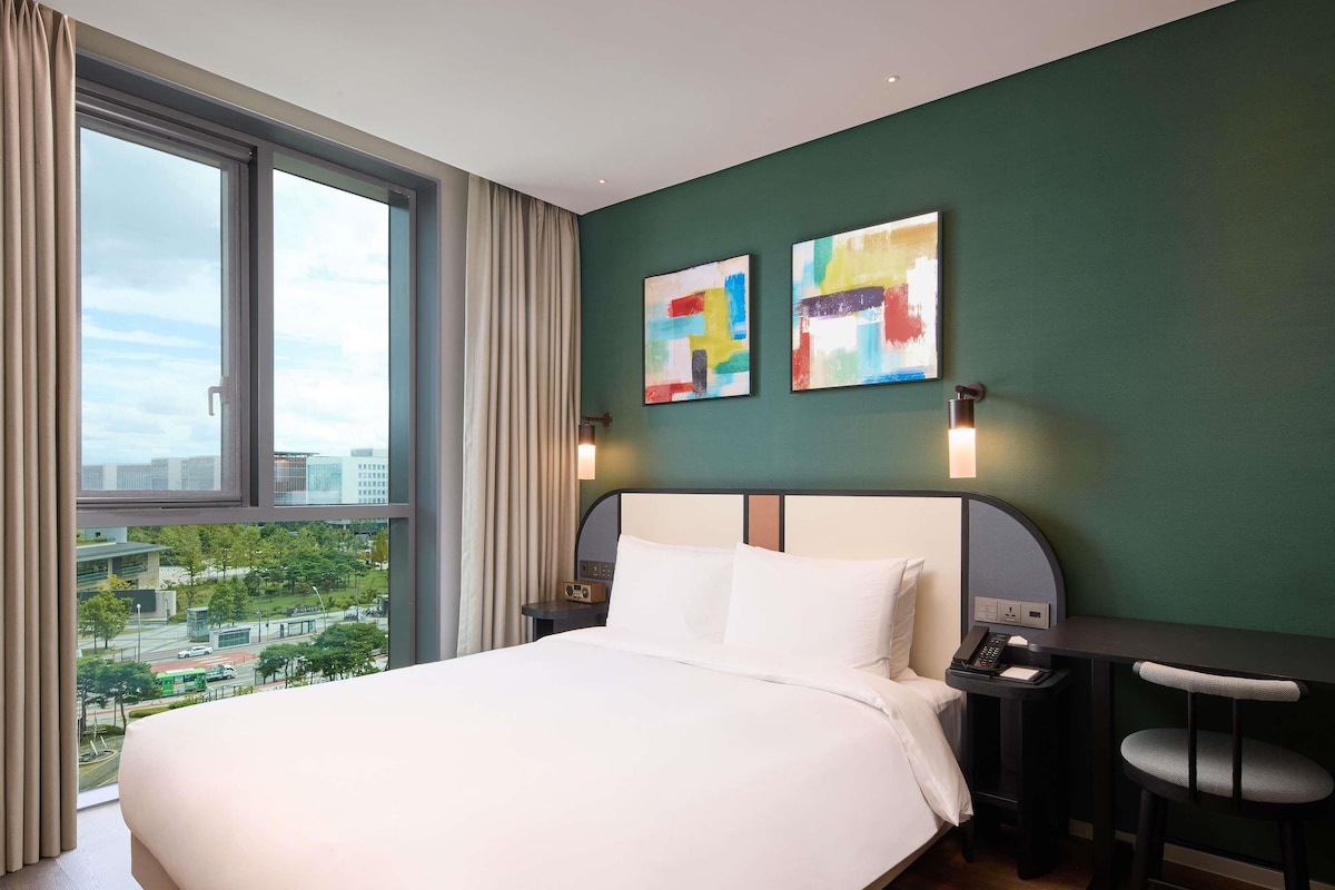A hotel room with a large bed, a desk, two chairs, a view of the city, and abstract art on the wall.