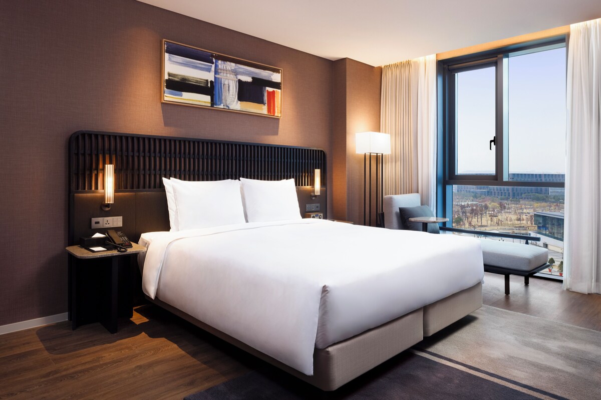Un moderno cuarto de hotel con una cama grande, una mesita de noche con teléfono, una lámpara de escritorio y vistas de la ciudad a través de un ventanal amplio.