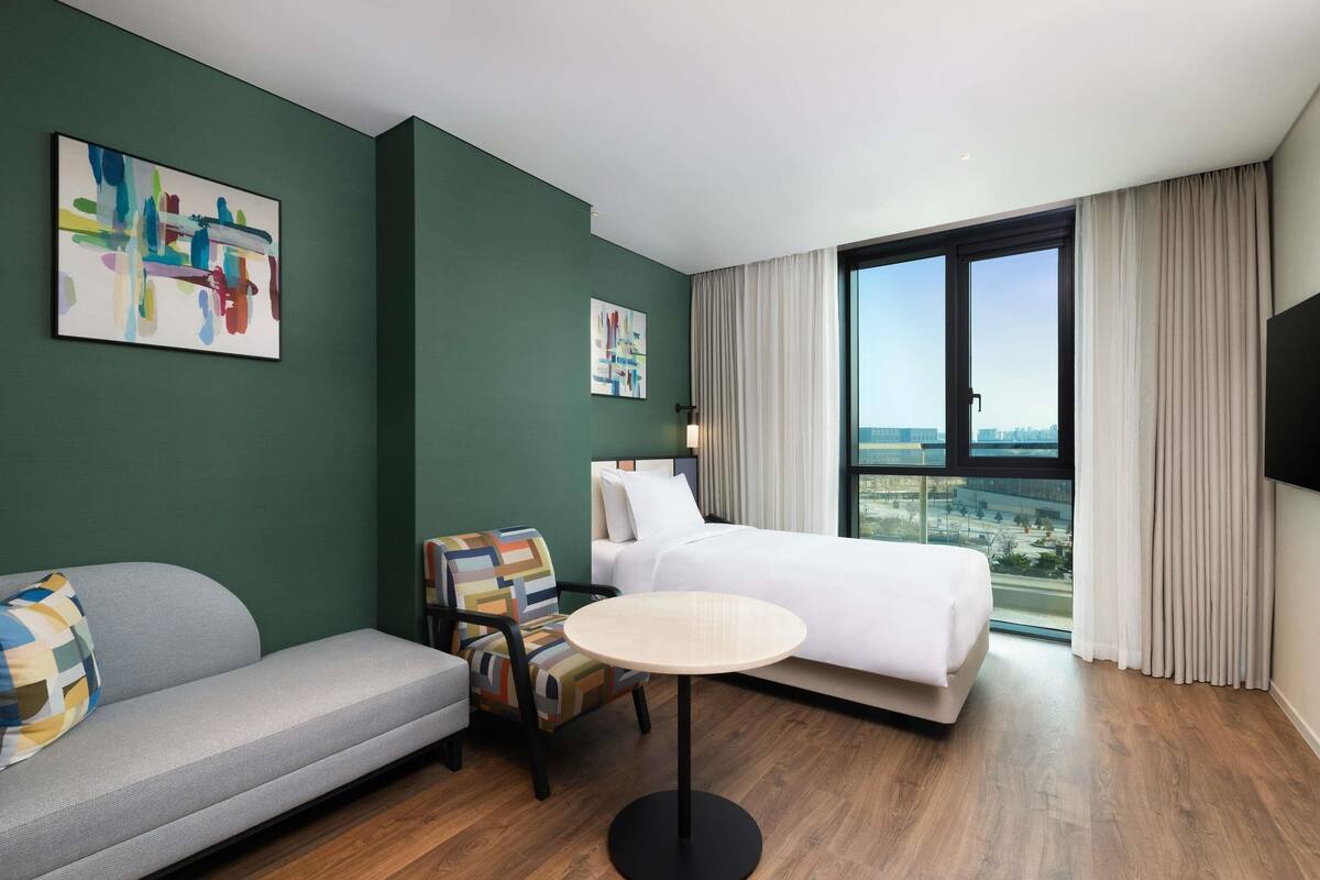 A hotel room with a bed, a sofa, a chair, a round table, and a large window with a city view.