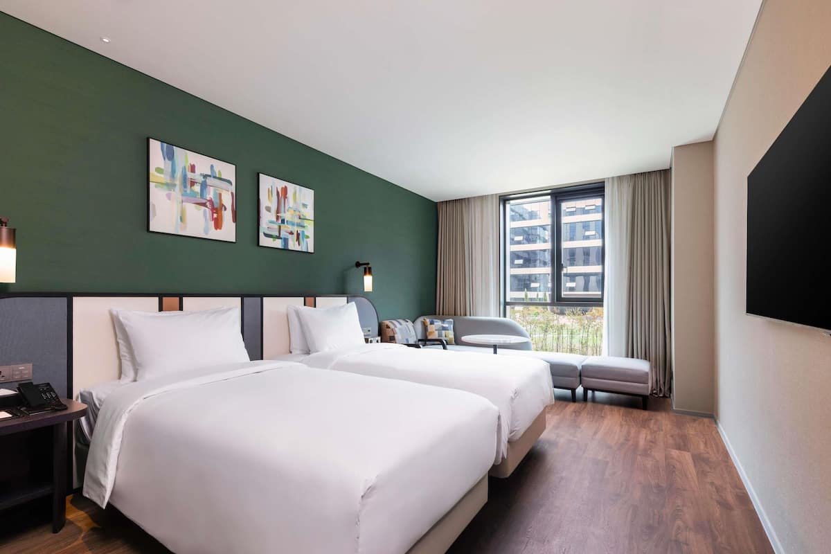 A hotel room with two beds, a desk, a chair, a TV, and artwork on the wall.