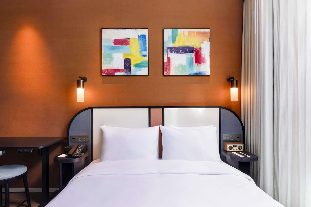 A hotel room with a bed, two bedside tables, a desk, and two abstract paintings on the wall.