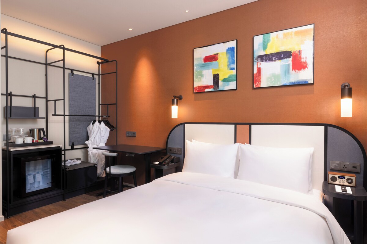 Una habitación de hotel con cama, escritorio, silla y un pequeño rincón de descanso. Hay dos obras de arte en la pared, un televisor y un minibar.