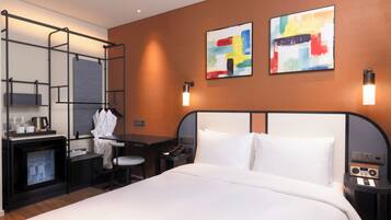 Una habitación de hotel con cama, escritorio, silla y un pequeño rincón de descanso. Hay dos obras de arte en la pared, un televisor y un minibar.