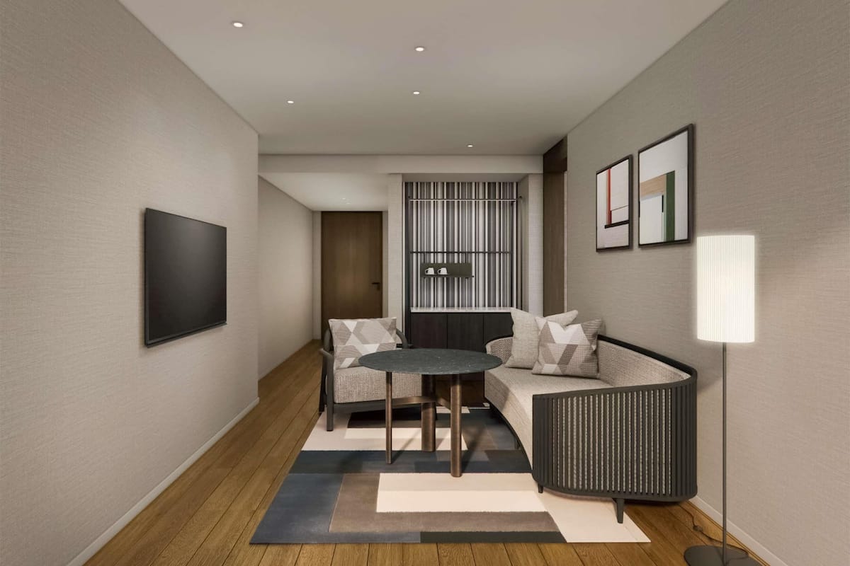 A modern hotel room with a sofa, a small round table, a floor lamp, and a TV mounted on the wall.