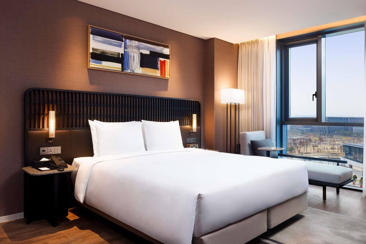 A modern hotel room with a large bed, a city view, a painting on the wall, and a desk with a lamp.