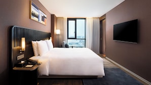 Une chambre d’hôtel moderne dotée d’un grand lit, d’un téléviseur à écran plat fixé au mur, d’une table de chevet avec une lampe et offrant une vue sur la ville par la fenêtre.