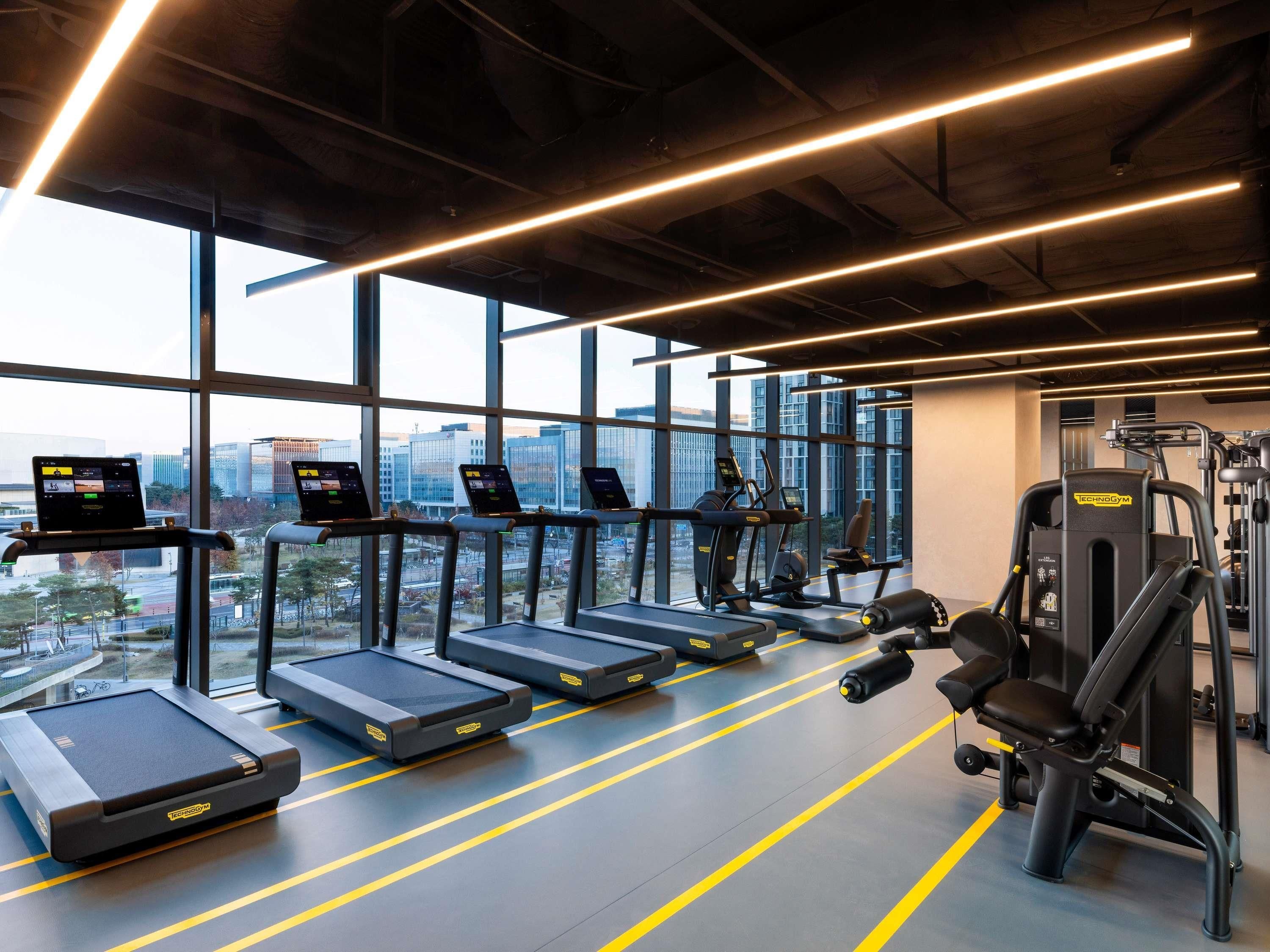 Une salle de sport équipée de tapis de course, de vélos d’exercice et d’appareils de musculation, de grandes fenêtres offrant une vue sur la ville et d’un éclairage moderne.