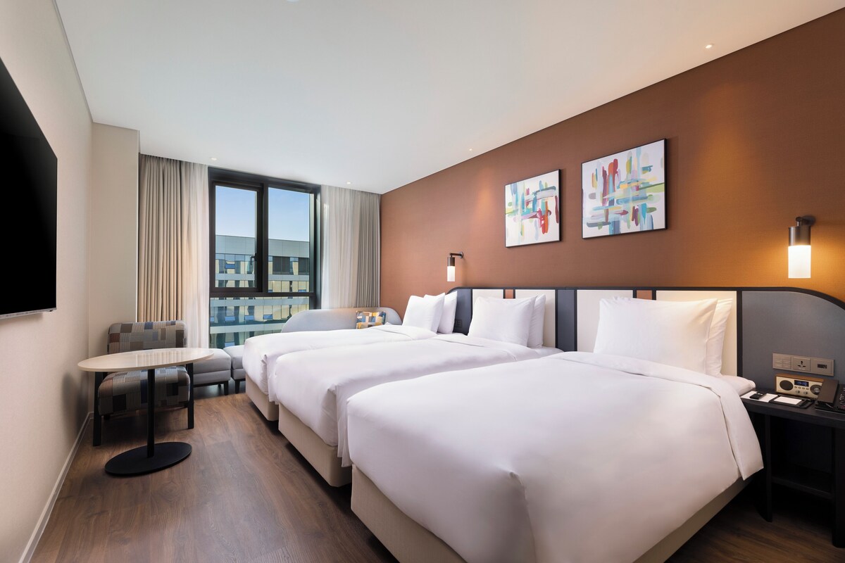 Habitación de hotel con dos camas, una mesita, una silla, televisión y obras de arte en la pared.