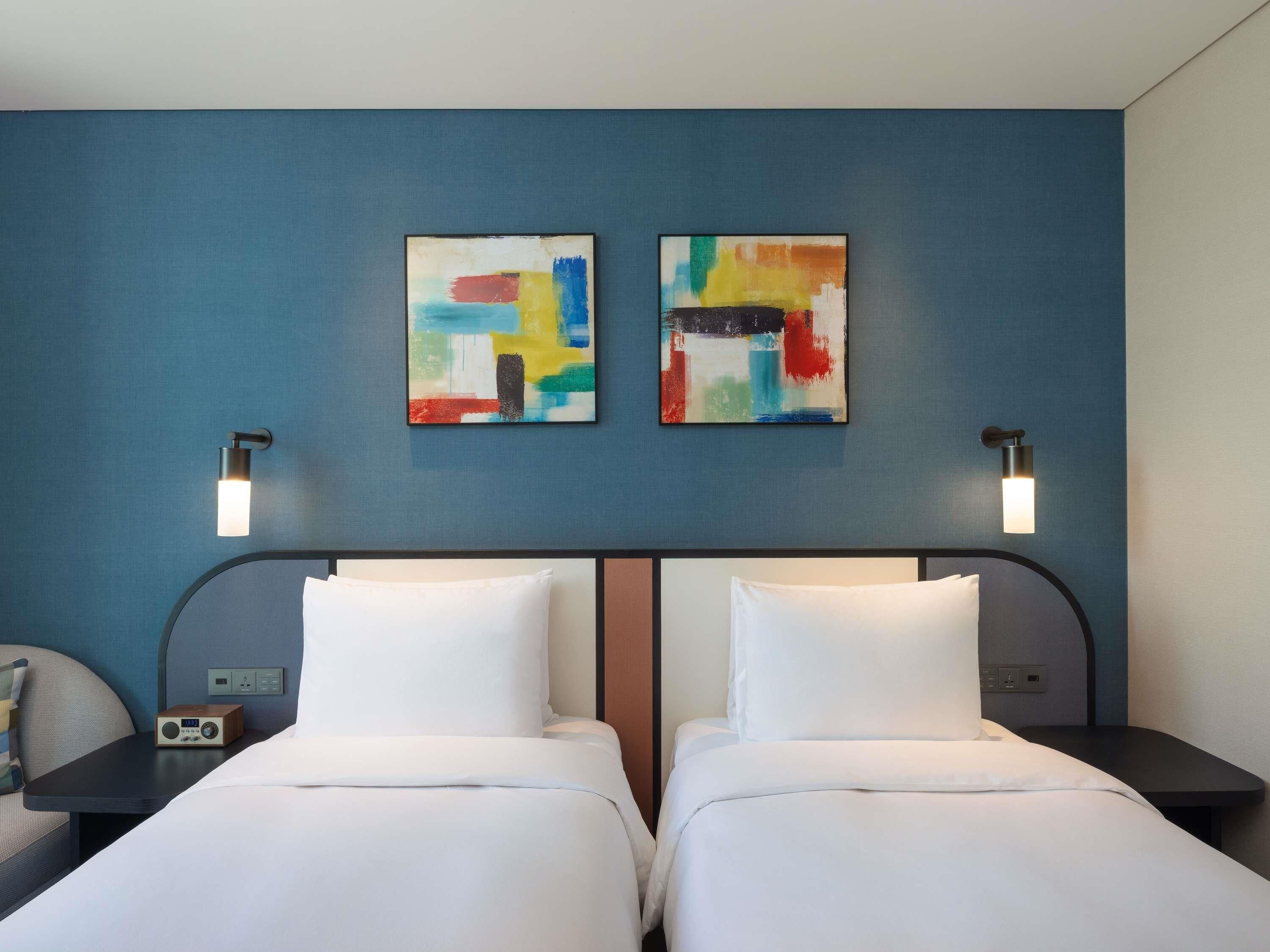 Quarto de hotel com duas camas, uma mesa de cabeceira, um rádio e duas pinturas abstratas na parede.