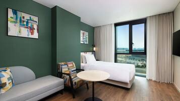 A hotel room with a bed, a sofa, a chair, a round table, and a large window with a city view.