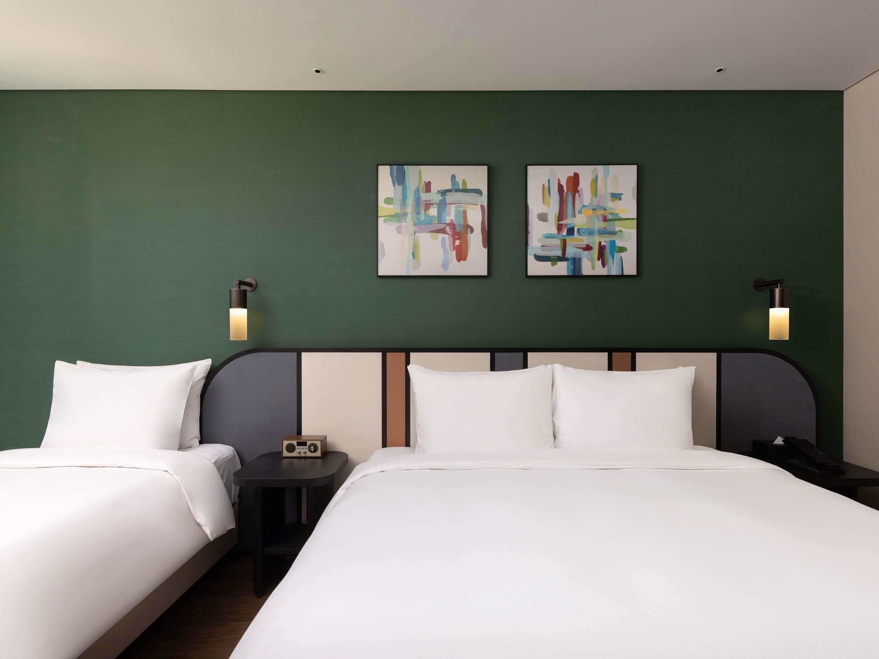 A hotel room with two beds, a headboard, two wall-mounted light fixtures, and two abstract paintings on the wall.