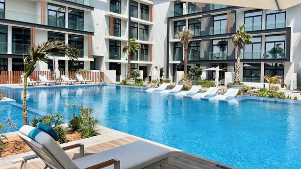 A modern indoor pool area with multiple lounge chairs and palm trees, surrounded by large windows and a multi-story building. - Waves - Ideal Dubai Retreat Elegant St (Dubai)