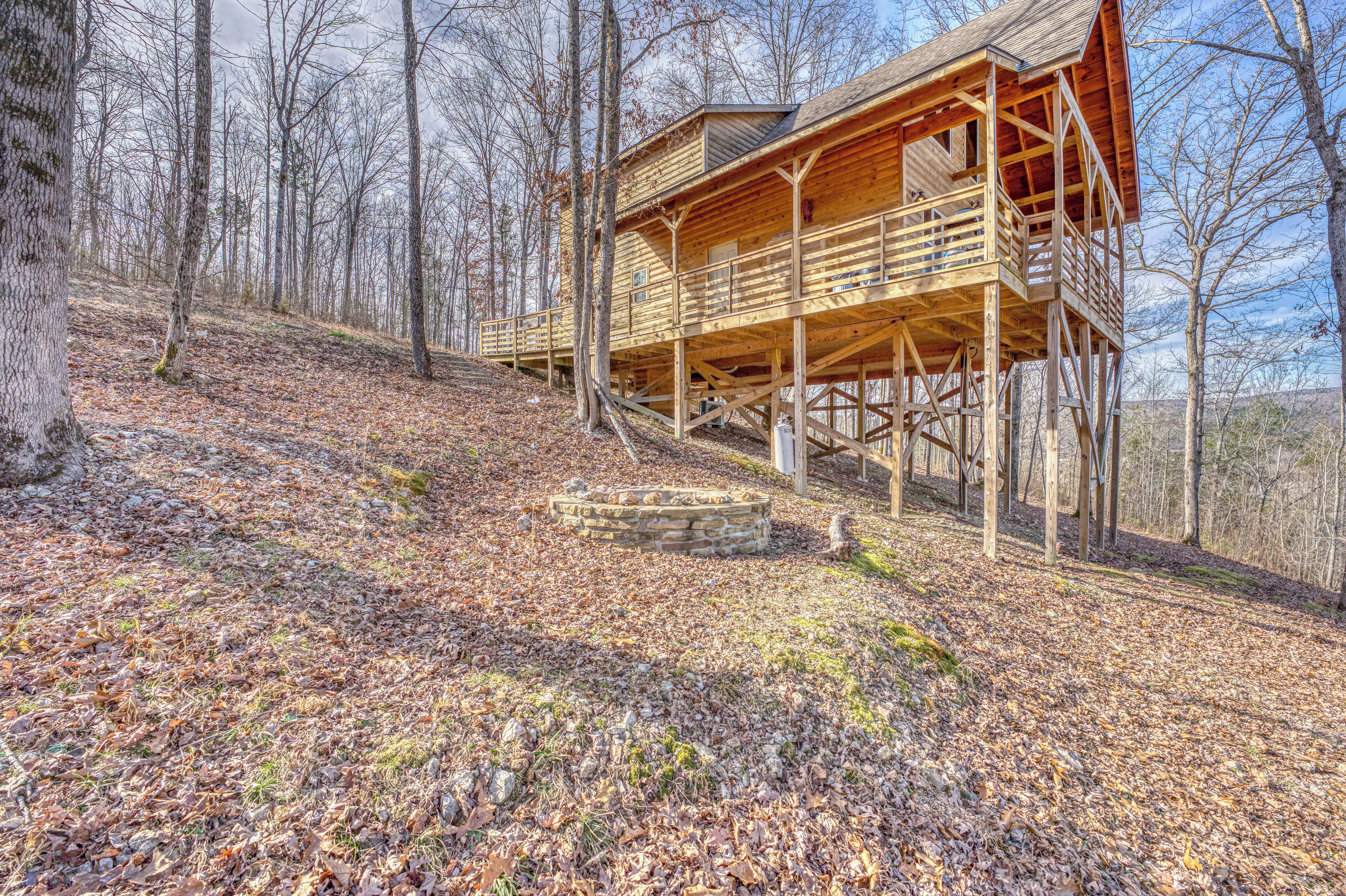A wooden cabin with a balcony, situated on a hillside with a stone fire pit in front.