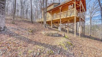 A wooden cabin with a balcony, situated on a hillside with a stone fire pit in front.