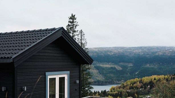 A dark wooden cabin with a white door and windows, a wooden deck, and a view of a lake and forested hills. - Amazing ski in/ski out Chalet in Åre - Övre Sadelvägen Ski Lodge   (Jämtlands län)