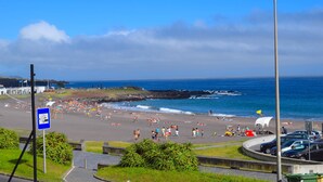 A beach with people sunbathing and swimming, a parking area with cars, and a clear blue sky.