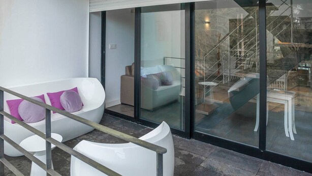 Un balcón moderno con mobiliario blanco, un ventanal que da a un espacio exterior, y vistas de una escalera y muebles de exterior.