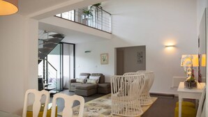 Amplia sala de estar con techo alto, escalera, conjunto de sofás, mesa de comedor con sillas y una pequeña mesa con lámpara.