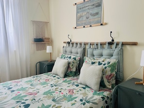Une chambre à coucher avec un lit, deux tables de chevet, une décoration murale en macramé, une carte encadrée et une fenêtre avec des rideaux.