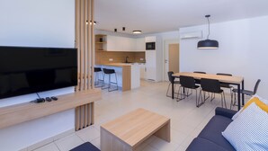 Ein modernes Wohnzimmer mit Flachbildfernseher, Couchtisch aus Holz und einer Couch. Es gibt eine Küchenzeile mit weißen Schränken und einem Esstisch mit Stühlen.