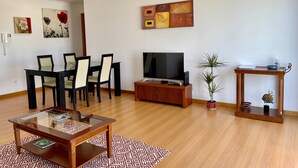 A living room with a glass-top coffee table, a TV on a wooden stand, a dining table with chairs, and a small side table with a plant and decorative items. - OCEAN TERRACE HOUSE by Azores HomeMade (Ribeira Grande)