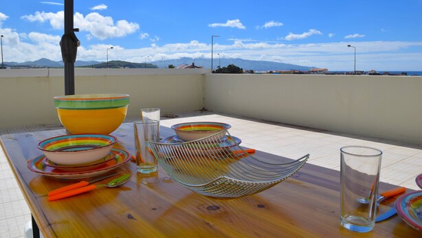 A dining table with colorful bowls, a glass, and cutlery on a terrace with a clear view of the sky and distant mountains. - OCEAN TERRACE HOUSE by Azores HomeMade (Ribeira Grande)