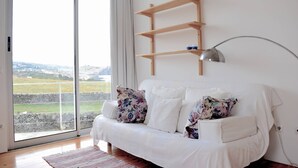 Un sofá blanco con cojines estampados, un estante de madera y una lámpara de pie en una habitación con un ventanal que ofrece vistas a un paisaje.