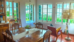 A dining area with a wooden table, chairs, and a bowl of fruit. There are large windows with a view of greenery outside. - QUINTA DAS BOLAS by Azores HomeMade (Ponta Delgada)
