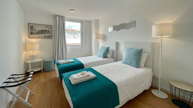 Une chambre d’hôtel avec deux lits, une petite table, une chaise, une fenêtre avec des rideaux, un tableau et une lampe sur pied.