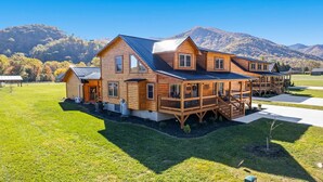 A large log cabin with multiple decks and a covered porch, surrounded by a well-maintained lawn and mountains in the background. - Meadow Breeze/360-Degree Mountain Views/Fire Pit (Maggie Valley)