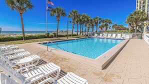 Une piscine entourée d’une clôture et de bâtiments, avec des chaises longues blanches et des palmiers.