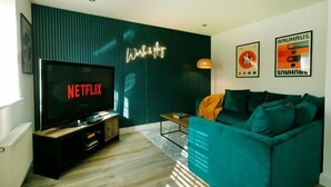 Una sala de estar moderna con un televisor de pantalla plana grande mostrando 'NETFLIX', un sofá color turquesa y un letrero de neón en la pared que dice 'Work & Play'.