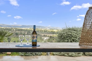 A bottle of wine and two glasses on a wooden table with a scenic view of hills and a clear sky. - Villa Cappero With Pool by Wonderful Italy (Noto)