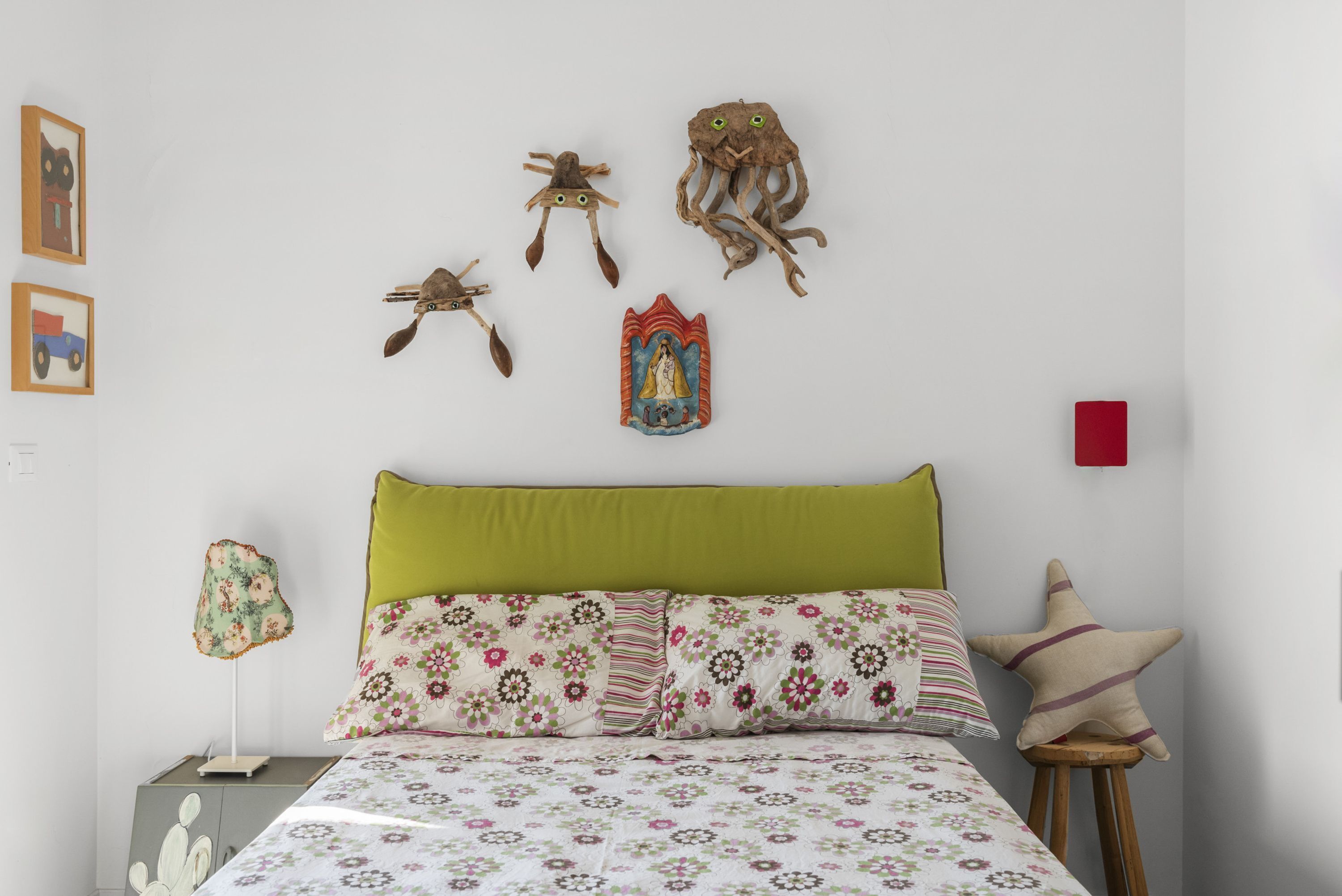 A bedroom with a green headboard, floral bedding, and wall art including a cat, octopus, and a religious icon.