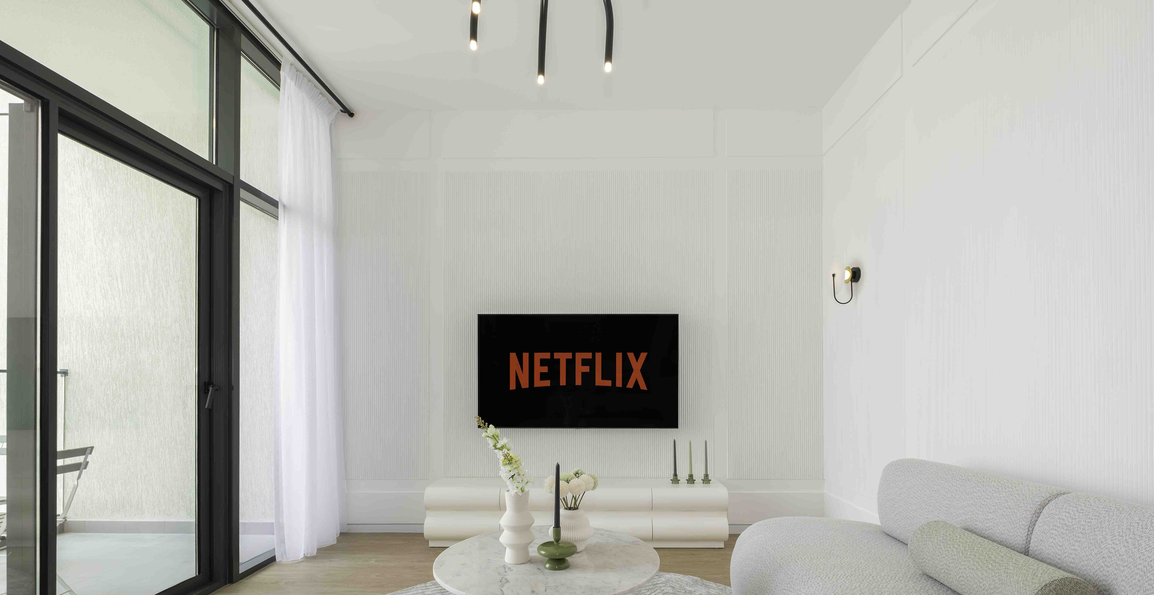 A modern living room with a large flat-screen TV displaying "NETFLIX," a white sofa, a coffee table with a vase of flowers, and a sliding glass door leading to a balcony.
