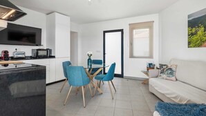 Cuisine moderne équipée d’un téléviseur à écran plat, d’un four à micro-ondes et d’un grille-pain. Un coin repas est aménagé avec une table en verre et des chaises bleues. Le salon comprend un canapé et un tapis bleu moelleux.