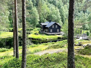 A dark-colored cabin with a deck, surrounded by lush greenery and a pond. - 10 Person Holiday Home in Kvinesdal (Kvinesdal)