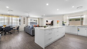 A modern kitchen with white cabinetry, a central island, and a dining area with a gray sofa and chairs. - Air Conditioning *  Secured Parking * BBQ * No Linen (South West Rocks)