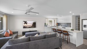 A modern living room with a grey sofa, a dining area with bar stools, and a kitchen with white cabinets. - Air Conditioning *  Secured Parking * BBQ * No Linen (South West Rocks)