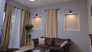 A living room with a brown leather sofa, two wall-mounted light fixtures, and a potted plant. - Najjera, Kampala 1bed With Balcony, Wifi & Parking (Kampala)