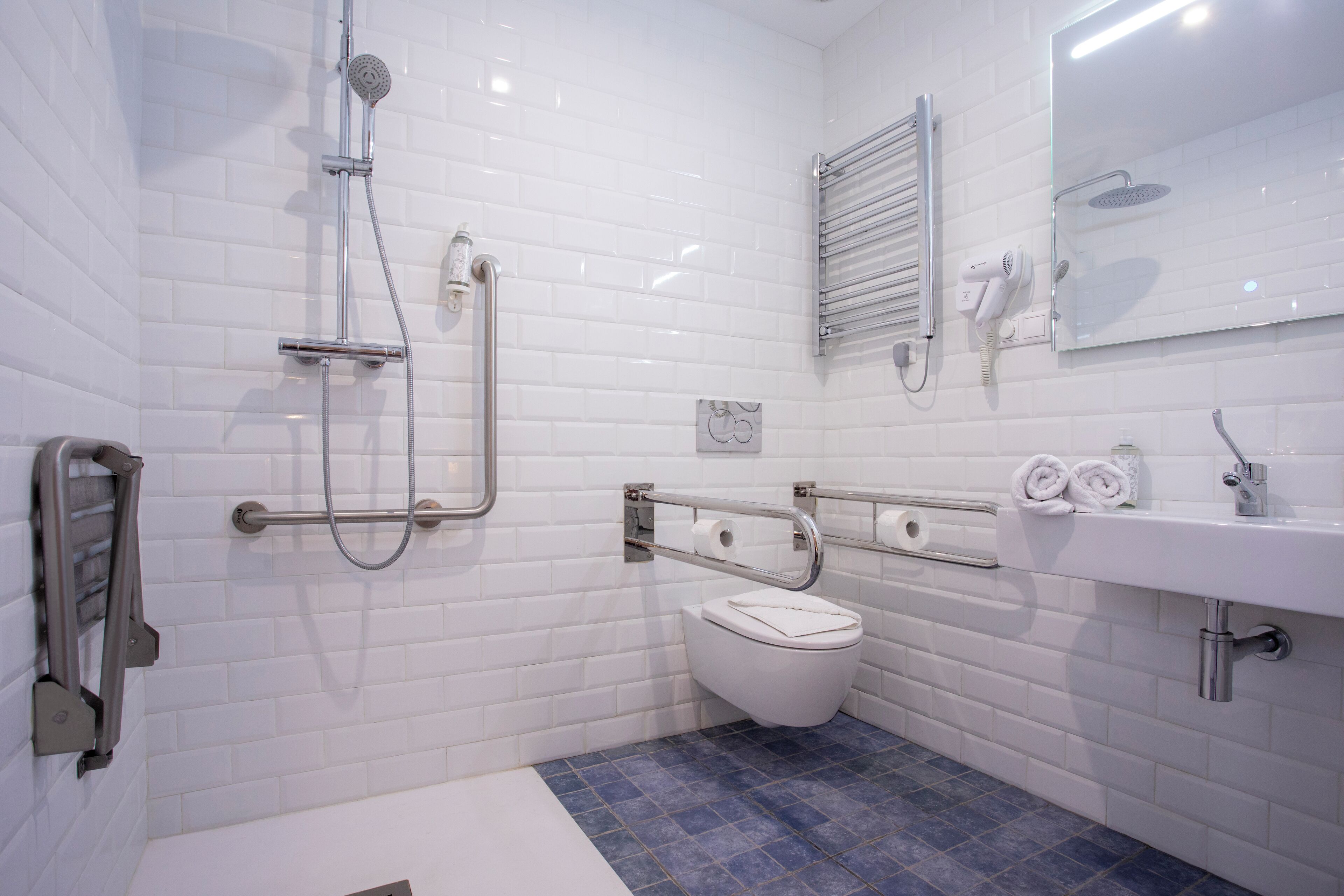 A modern bathroom with white tiled walls, a blue-tiled floor, a white toilet, a sink with a mirror, and a shower with a handheld showerhead.