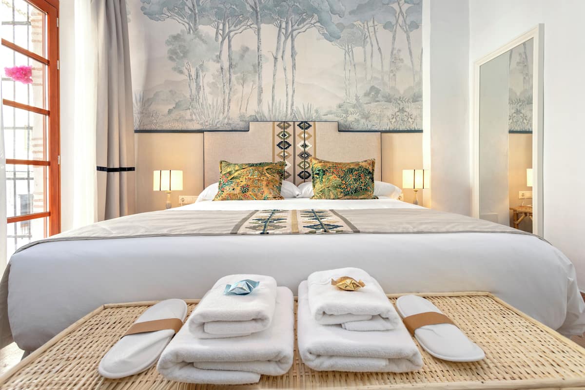 Habitación de hotel con una cama grande, dos almohadas, un cabecero con panel decorativo, una bandeja de mimbre con toallas y un ventanal con vistas a árboles.
